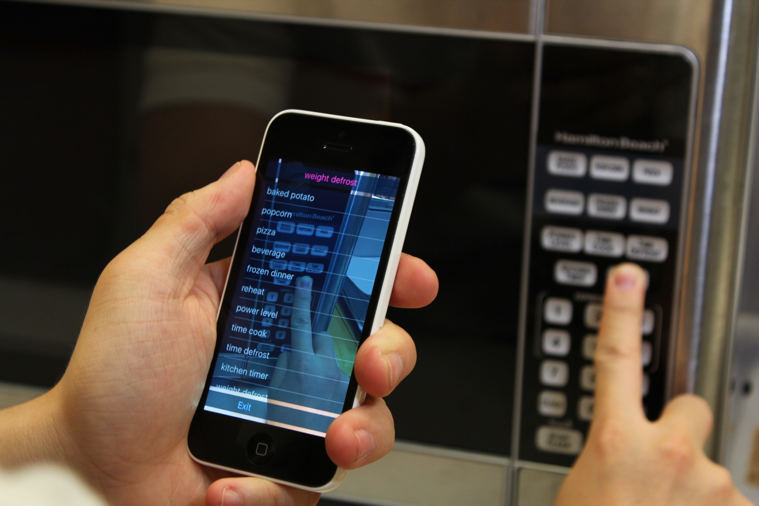 The user is holding the phone in portrait mode with one hand, and aiming the camera towards an inaccessible microwave control panel. The user‚Äôs other hand is exploring on the panel. The VizLens iOS app is providing audio feedback and guidance to the user.