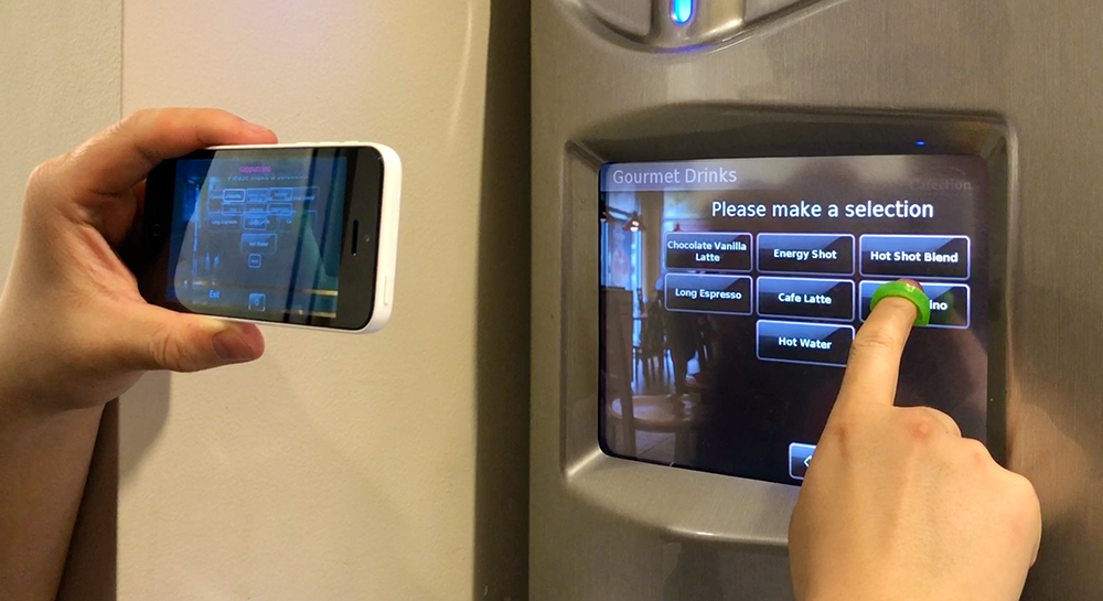 The user is holding the phone in landscape mode with one hand, and aiming the camera towards a touchscreen coffee machine. The user‚Äôs other hand is wearing a fingercap exploring on the screen. The StateLens iOS app is providing audio guidance to the user.